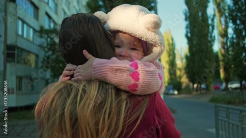 A child in a cozy hat hugs a parent tightly as they walk in a park under clear skies. Bright autumn trees surround them, adding warmth to the moment.