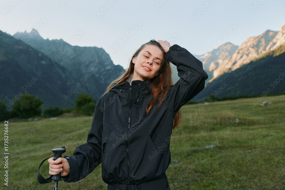 Naklejka premium A cheerful hiker stands in a scenic mountain meadow wearing a black jacket, leaning on a trekking pole, with a confident smile as sunlit peaks rise behind her.