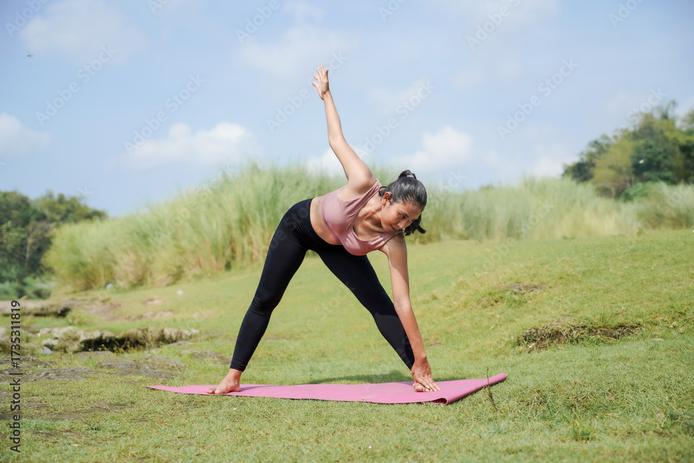 Fototapeta premium Mental and physical health. A slender, tanned Asian girl in sportswear does yoga by a calm river in the morning. The woman stands in Extended Triangle Pose with her arms down and up.