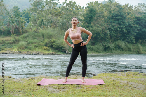 A slender Asian girl with brown skin in sportswear is doing hip circle stretching movements in the open air on the edge of a beautiful river, before doing sports.