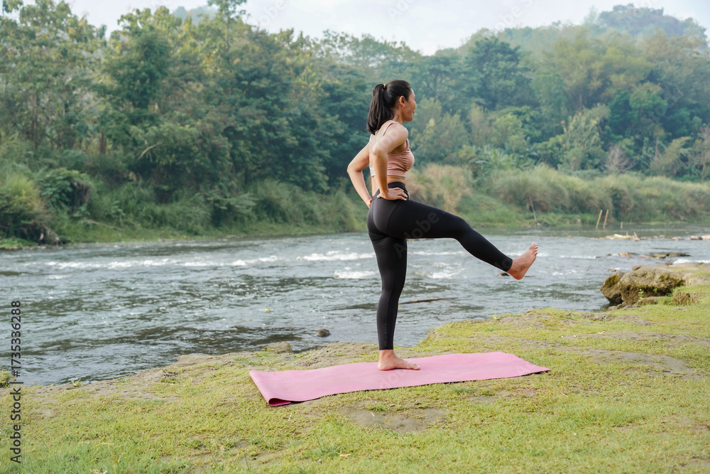 Fototapeta premium A slender Asian girl with brown skin in sportswear doing stretching movements and swinging her legs in the open air on the edge of a beautiful river, before doing sports.