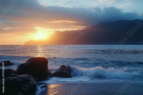 Sunset over the ocean with gentle waves crashing on dark rocks and silhouetted mountains under a partly cloudy sky