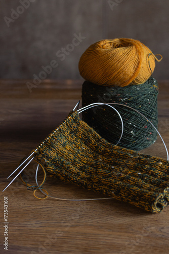Cozy knitted socks with yarn ball and knitting needles. Knitting process of wool socks. Cozy autumn vibes. Yellow and green tweed yarn on wooden table. Knitting hobby. Knitwear concept. 
