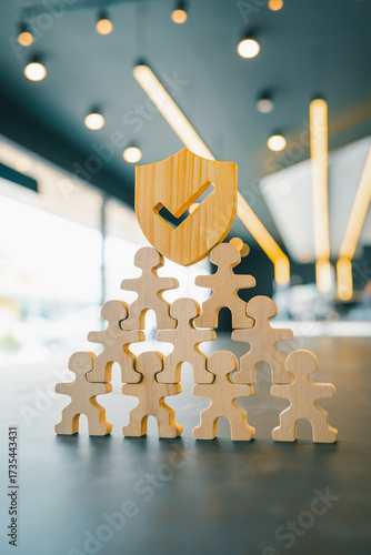 Wooden human pyramid with shield and check mark representing teamwork, trust, insurance protection, risk management, and corporate security concept for business, HR, or assurance services.