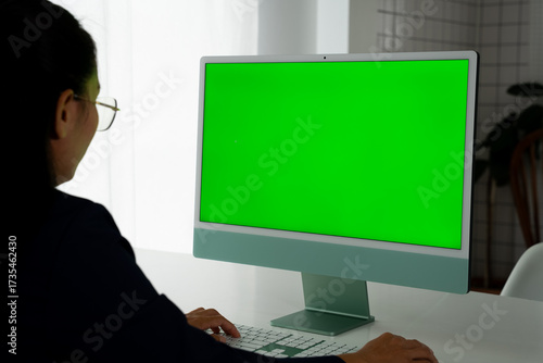 Asian Female Engineer Engaging with Desktop Computer Featuring Green Chroma Key Screen. Modern Office Setting for Engineering and Creative Tasks, Emphasizing Technology and Innovation.