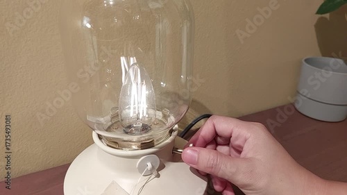 Close-up video of a person’s hand turning on a modern table lamp with a glass shade, emitting warm cozy light. Perfect for illustrating home interior, night routine, electricity, lighting