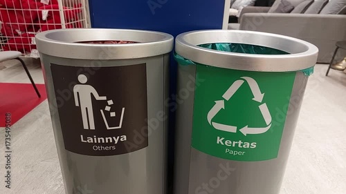 Video of recycling and waste bins placed side by side indoors. One bin is labeled for paper recycling with a green symbol, while the other is marked for general waste. The scene promotes environmental