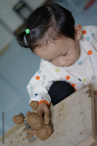 A young child focused on playing with clay, engaged in the process of creation. The child is seated at a wooden table, molding the clay with focus and care