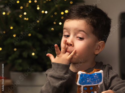 Little boy near a decorated Christmas tree holds a fake ice cream shaped like a Santa boot or gingerbread. Child in pajamas tastes sweets and licks fingers in cozy dim light festive New Year atmospher