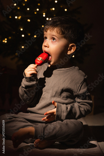 Little boy near a decorated Christmas tree holds a fake ice cream shaped like a Santa boot or gingerbread. Child in pajamas tastes sweets and licks fingers in cozy dim light festive New Year atmospher