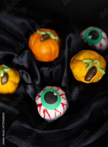 Halloween pastries with small orange and yellow pumpkins and spooky eyeball sweets with red veins. Woman dressed as a witch bites or holds dessert near her eyes on dark textile background.