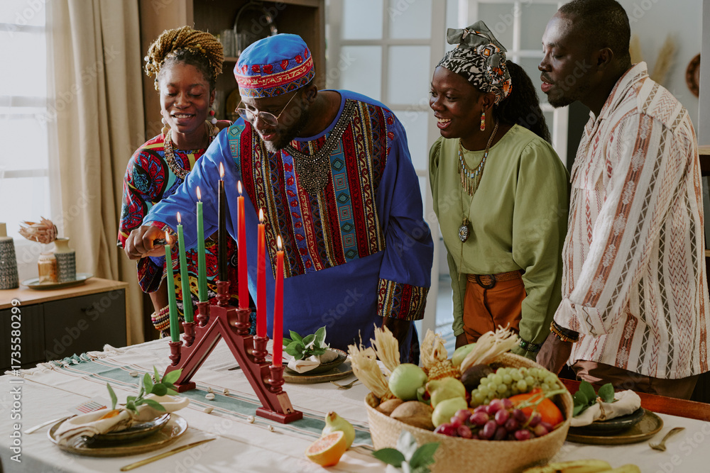 Fototapeta premium Group of Black adults and young adult woman celebrating Kwanzaa, lighting kinara candles together at table with traditional fruits and decorations, smiling and interacting