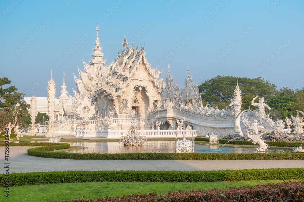 Fototapeta premium White temple Wat Rong Khun with intricate carvings and bridge over water surrounded by lush garden at sunrise, Chiang Rai, Thailand. Buddhist temple in Northern Thailand
