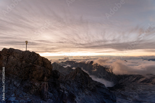 Silhouette of the Zugspitze summit with cross at dusk, dramatic mountain ridge above a sea of clouds under moody evening sky