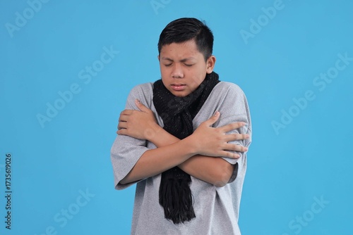 Young Boy Shivering and Feeling Cold, Hugging Himself Tightly While Wearing Scarf on Blue Studio