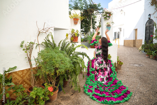 Young, pretty, brunette, flamenco dancer, doing dancing poses with her arms, showing off her beautiful dress, sitting on a chair, next to a well in a nice, typical Andalusian courtyard.