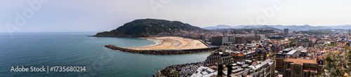 View of the vast expanse of the blue sea contrasts sharply with the golden sandy beach, teeming with tiny figures, embraced by a sturdy rock barrier, San Sebastian, Donostia, Spain.