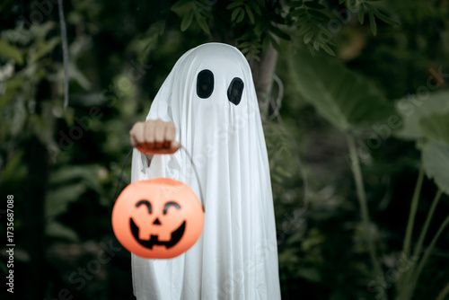 A young boy dressed as a ghost for Halloween wears a white sheet with black eye holes. Holding a pumpkin jack-o’-lantern bucket, he stands outdoors surrounded by dark green foliage