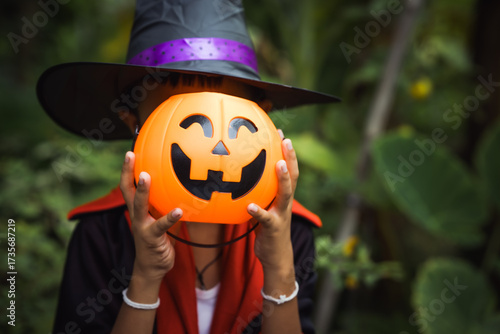 Two young children in Halloween costumes stand outdoors, holding pumpkin buckets with jack-o’-lantern faces. Wearing witch hats festive outfits, they share playful smiles, surrounded by lush green