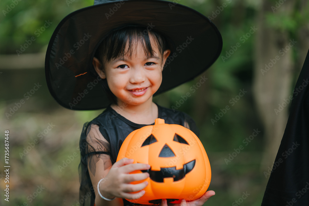 Fototapeta premium A cute little girl smiles brightly, dressed as a witch for Halloween. She wears a black pointed hat, an orange-and-black costume, holding a jack-o’-lantern pumpkin bucket outdoors among green trees
