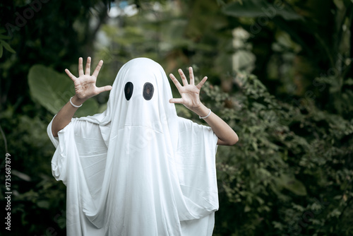 A young boy dressed as a ghost for Halloween wears a white sheet with two black eye holes. He raises both arms in a spooky gesture, surrounded by dark green foliage
