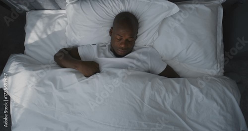 Man Lies in Bed, Stretches, Slowly Wakes Up in Morning Light. Male African American Looks Sleepy, Reluctantly Wakes Up, Resting on White Pillows. Concept of Waking Up, Morning Routine, Sleep Inertia.