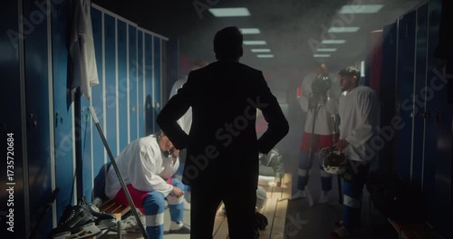 Frustrated Ice Hockey Coach Standing with His Back to Camera, Shouting at His Team in a Locker Room After a Loss. Players Sit Quietly, Looking Down as They Endure His Harsh Criticism
