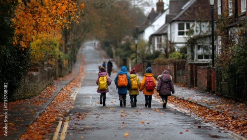 Children walking down a tree-lined street in autumn, with colorful leaves on the ground and houses in the background.