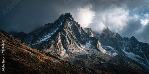 Dramatic sharp mountain peaks under moody sky, with sunlight breaking through clouds, highlighting rugged terrain and snow dusted slopes, creating breathtaking scene