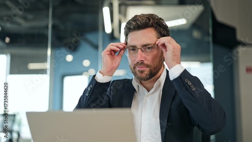 Handsome businessman dressed in a white shirt and suit jacket works on a laptop in a modern office. Professional male smiles, feeling happy and successful about his work.
