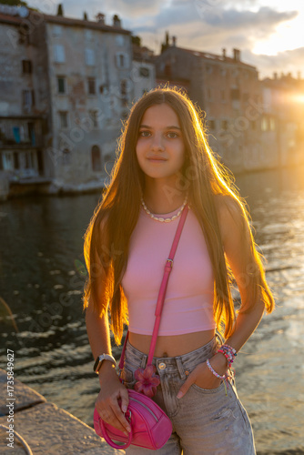 Teen girl posing by canal at sunset. Stylish teenage girl in pink outfit posing near waterfront at golden hour, with historic buildings and sun flare in background.
