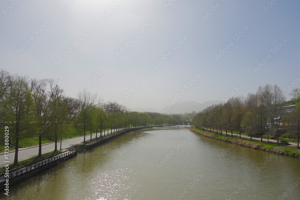 Fototapeta premium Wide, straight canal or river running between tree-lined roads with distant hazy mountains under a bright, misty sky. Spring landscape scenery.