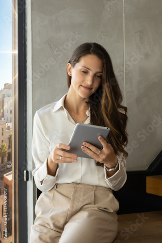 A professional woman in a white blouse and beige trousers stands by a large office window, holding and using a tablet