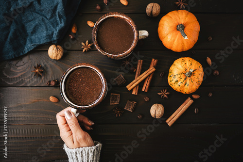 Top view of female hand in sweater holding mug of hot chocolate, pumpkins and spices on rustic wooden background, cozy autumn scene