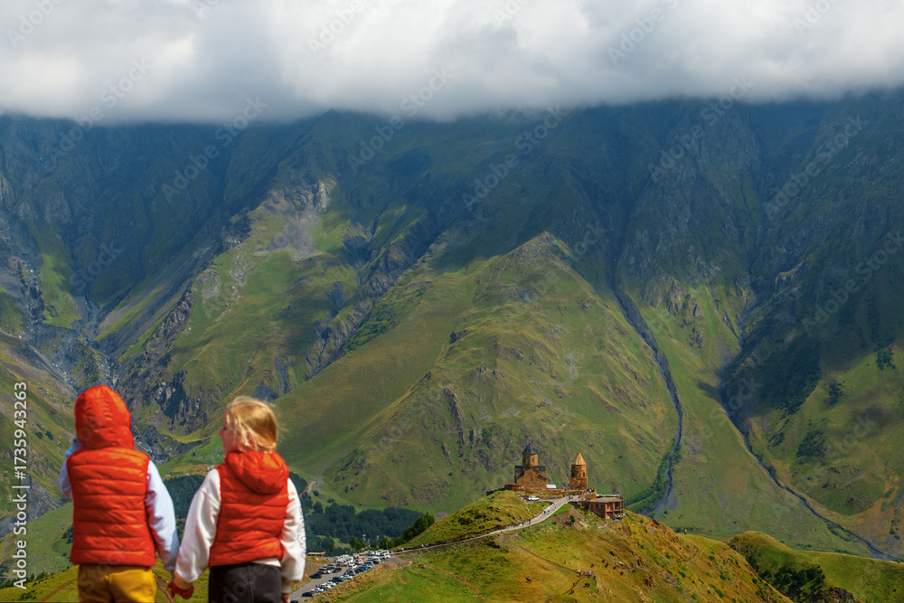 Fototapeta premium Two young girls holding hands and looking at the famous Gergeti Trinity Church in Georgia with many tourists around, capturing a moment of travel, culture, and heritage in the scenic mountains.