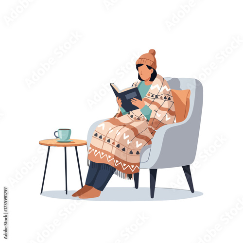 Woman wrapped in blanket sits in chair reading book with cup tea on side table