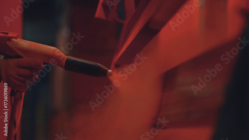 Worker's gloved hand operates an electrostatic spray gun, applying a vibrant red powder coating onto a metal surface in an industrial manufacturing environment, ensuring durable protective finish