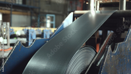 Steel sheet metal unrolling from a large coil, moving through the guiding rollers of industrial machinery in a modern manufacturing factory environment