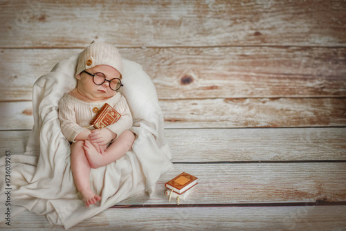 A newborn baby. The baby is 10 days old. The boy wears glasses. The baby is dressed in a beige bodysuit and a hat, and he is sitting in a chair. The newborn boy is holding a book. Sleeping. Smart.