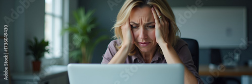 A worried middle-aged woman in a corporate office displays fatigue while staring at her laptop. The image accentuates stress in the workplace, providing room for text placement.