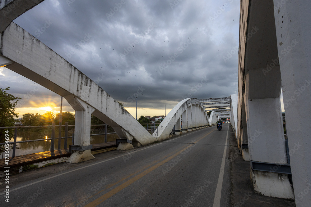 Obraz premium Dramatic arch bridge spanning serene river, illuminated by sunset light piercing through cloudy, expressive sky. Robust concrete structure stands against atmospheric weather, creating peaceful