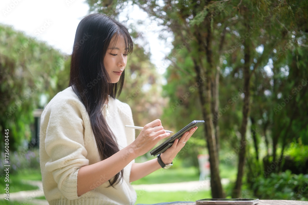 Obraz premium Focused woman sketching on digital tablet with stylus in lush outdoor environment.
