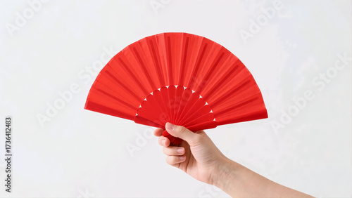 Close-up of a hand holding a vibrant red fan against a clean white background. The image highlights the texture and color of the fan, creating a simple yet striking composition.