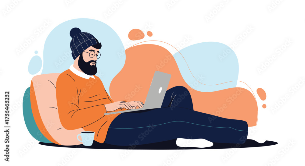 Fototapeta premium A man with a beard wearing a beanie and an orange sweater sits on a couch and works on a laptop with a cup of coffee nearby.