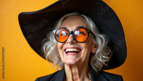 Funny elderly woman with silver hair wearing oversized black witch hat and orange glasses laughing joyfully on bright background halloween portrait