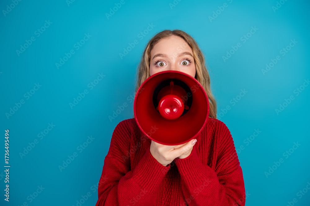 Fototapeta premium Cheerful young woman in casual attire shouting excitedly through a red megaphone against a vibrant blue background