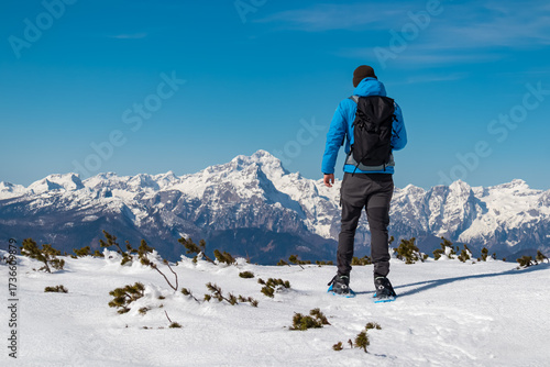 Hiker in blue jacket stands on snowy Bärentaler Kotschna plateau, gazing towards the towering Triglav and dramatic Karawanks range, capturing the spirit of alpine adventure on Austria-Slovenia border