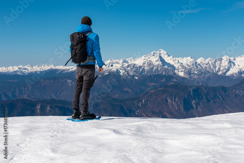 A hiker in blue winter gear stands on snowy ground, gazing toward the distant Julian Alps and Triglav from Bärentaler Kotschna in the Karawanks, surrounded by dramatic alpine scenery and clear skies.