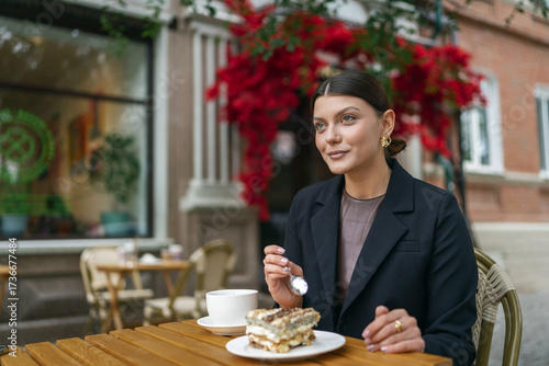 Joyful woman at an outdoor cafe celebrating with a slice of cake featuring a single birthday candle. She wears a smart black blazer and has a cup of tea on the wooden table.