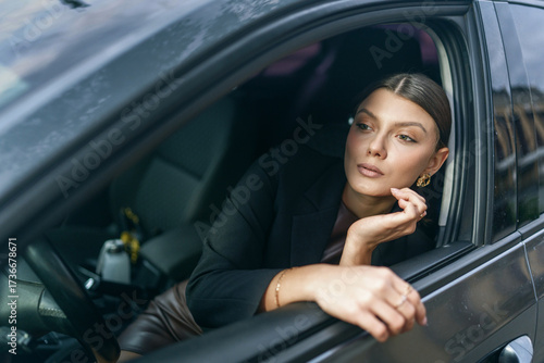 Stylish woman in a black blazer looks out from a car window. The portrait captures her sophisticated style and confident gaze against an urban background.